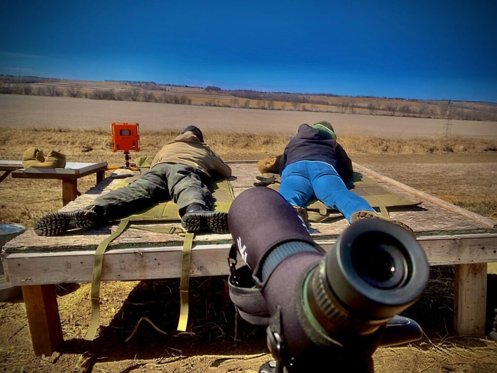 A picture of two men lay on a platform with their rifles pointed away from the camera. One in a blue tee shirt another in camo both with their baseball caps backwards focused on the targets down range. In the foreground a spotters scope looks over their shoulder.