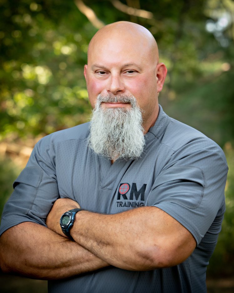 Paul is a tall man with broad shoulders, a bald head, and a silver well groomed beard. He stands in a gray RAM Mast Training T Shirt