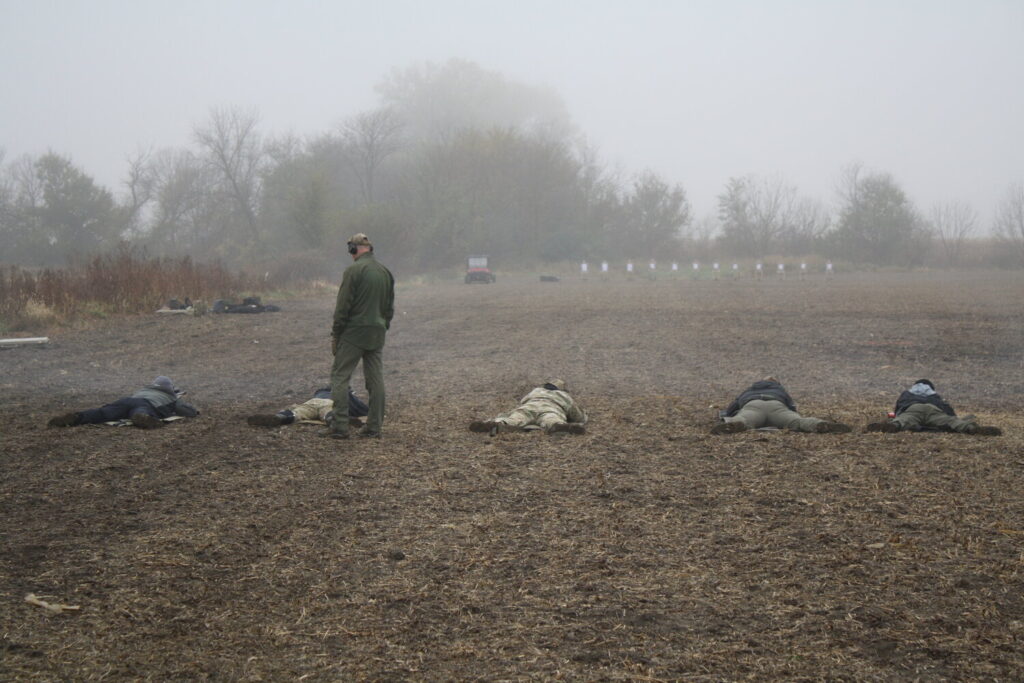 Five men in a muddy field cast in a light fog as an instructor supervises. White Silhouette targets sit off in the distance