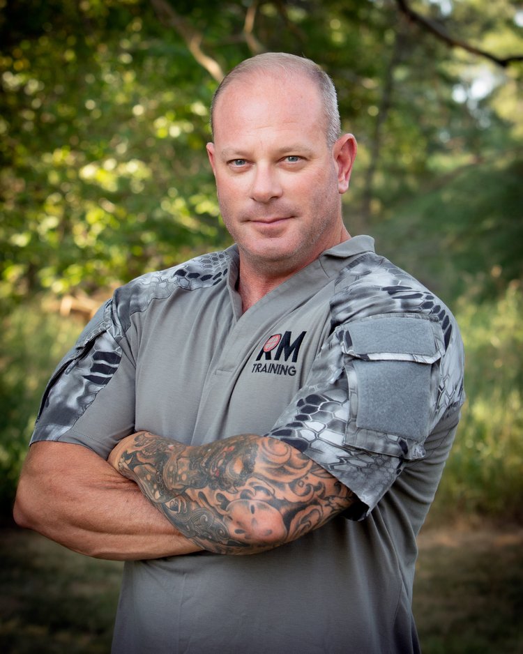 Instructor Tim is a tall man withs square shoulders  with a short receding hairline. Tim is standing with his arms crossed across his chest showing off his large forearm muscles and tattoo sleeve. 